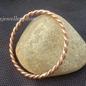 Könnte beinhalten: Ein kupferfarbenes, verdrehtes Armreif-Armband liegt auf einem glatten, hellfarbigen Stein. Das Armband hat ein geflochtenes Design und ist vor einem dunkelblauen, strukturierten Hintergrund platziert. Der Text "Luckyjeweller store" ist sichtbar.