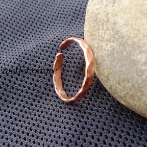 Può includere: Un anello color rame con un design aperto e una superficie martellata e testurizzata. La fascia presenta una forma leggermente irregolare e organica con una finitura metallica, mostrando una distinta tonalità bruno-rossastra.