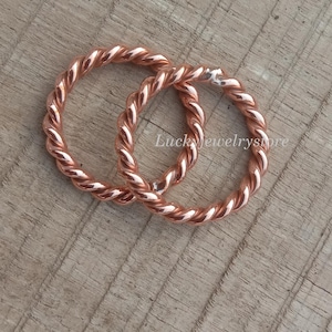 May include: Two copper-colored, twisted metal rings, possibly jewelry, are displayed on a wooden surface. The rings are circular with a braided design. The background is a light brown wood grain.