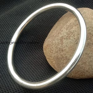 Pode incluir: Uma pulseira tipo bracelete de tom prateado, lisa e polida, com um design circular simples. A superfície metálica é reflexiva e de espessura uniforme, formando um aro contínuo. O texto "luckyjewellerstore" é visível no lado esquerdo.