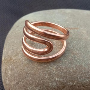Könnte beinhalten: Ein kupferfarbener Ring mit Spiraldesign. Der Ring besteht aus drei verbundenen Bändern, die sich krümmen und überlappen, wodurch eine einzigartige Form entsteht. Der Ring wird auf einem glatten, grauen Stein präsentiert.