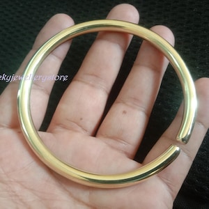 Może przedstawiać: Złota, otwarta bransoletka. Bransoletka ma prosty, gładki i zaokrąglony design. Tekst "Luckyjewelrystore" jest widoczny na zdjęciu. Bransoletka jest trzymana w dłoni.