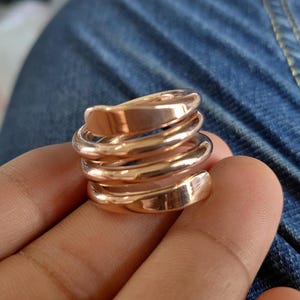 Könnte beinhalten: Ein polierter Ring in Roségold-Optik mit einem mehrschichtigen Spiraldesign. Das Band besteht aus vier durchgehenden, überlappenden Windungen, die sich um den Finger legen und eine breite, gestapelte Optik mit glatter, reflektierender Oberfläche erzeugen.