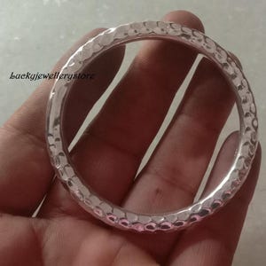 Könnte beinhalten: Ein kreisförmiger Armreif aus Silber mit einer gehämmerten, genoppten Textur auf der gesamten Oberfläche. Das Metall hat eine reflektierende, polierte Oberfläche. Der Text "Luckyjewellerystore" ist schwarz auf der linken Seite eingedruckt.