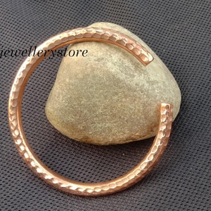 Könnte beinhalten: Ein kupferfarbenes Armband mit gehämmertem Finish liegt auf einem hellbraunen Stein vor einem dunkelgrauen, strukturierten Hintergrund. Das Armband hat ein offenes Design und eine strukturierte Oberfläche. Der Text "Luckyjewellerystore" ist sichtbar.