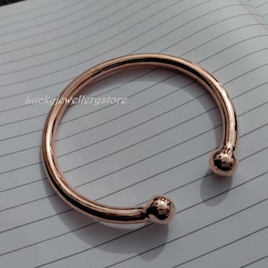 Könnte beinhalten: Ein roségoldenes Armreif-Armband mit glattem, abgerundetem Design. Die offenen Enden des Armbands weisen kleine, kugelförmige Akzente auf. Das Armband wird auf einer linierten Papieroberfläche präsentiert.