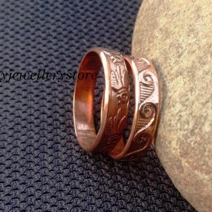 May include: Two copper rings with engraved designs. One ring features a bird motif, while the other displays a swirling wave pattern. The metallic bands have a warm, reddish-brown finish and are positioned upright next to a smooth, light-colored stone.