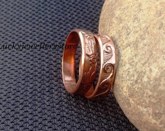 May include: Two copper rings with engraved designs. One ring features a bird motif, while the other displays a swirling wave pattern. The metallic bands have a warm, reddish-brown finish and are positioned upright next to a smooth, light-colored stone.