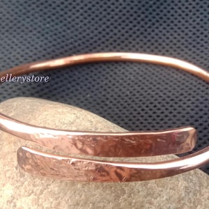 Puede incluir: Una pulsera de cobre con diseño abierto y textura martillada en los extremos superpuestos. El metal tiene un acabado pulido y un tono marrón rojizo cálido. El texto "kyjewellerystore" es visible en el lado izquierdo de la pulsera.