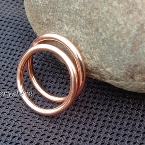 Könnte beinhalten: Ein doppelter Ring aus Roségold liegt auf einer dunkelgrauen, strukturierten Oberfläche neben einem glatten, grauen Stein. Der Ring besteht aus zwei verbundenen Bändern. Der Etsy-Shop-Name, luckyjewellerystore, ist unten links sichtbar.