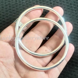 May include: Two smooth, polished silver-tone bangle bracelets with a rounded profile. The text Luckyjewellerystore is visible across the center of the image, overlaid on the metallic surface of the circular jewelry pieces.