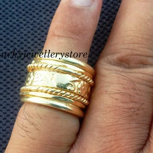 Könnte beinhalten: Ein goldfarbener Ring mit breitem Band, verziert mit aufwendigen geätzten Mustern und seilartigen Details. Der Ring wird an einem Finger getragen, wodurch sein Design und seine Handwerkskunst hervorgehoben werden. Die Oberfläche des Rings reflektiert das Licht.