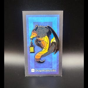May include: A black, yellow, and blue enamel pin of a dragon holding a lantern. The dragon is curled up and has a fluffy mane. The pin is on a blue background with a white border. The text "DragonfeathersArt" is on the bottom of the pin.