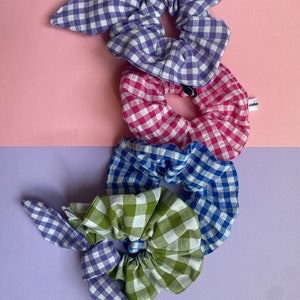 Puede incluir: Cuatro scrunchies de gingham en diferentes colores y patrones. Uno es un lazo de gingham morado y blanco, uno es un scrunchie de gingham rosa y blanco, uno es un scrunchie de gingham azul y blanco, y uno es un scrunchie de gingham verde y blanco.