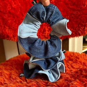 Peut inclure: Plusieurs chouchous en denim bleu clair et foncé. Les chouchous sont fabriqués à partir de tissu denim recyclé et présentent un design à volants. Les chouchous sont tenus par une main, et l'arrière-plan est une surface orange floue.
