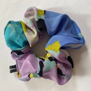 Puede incluir: Un scrunchie para el cabello colorido con un estampado abstracto morado, azul, turquesa, amarillo y negro. El scrunchie tiene una pequeña etiqueta negra con la palabra "ations." impresa en ella.
