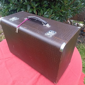 May include: A vintage brown suitcase with a textured finish, silver corner protectors, and a black handle. The suitcase is secured with two silver latches and a purple string. It rests on a red surface.