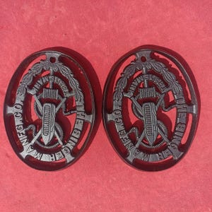 May include: Two oval-shaped black cast iron Singer Manufacturing Company trivets. Each piece features an intricate relief design of a sewing machine shuttle, crossed needles, and a laurel wreath border with the text THE SINGER MFG. CO. TRADE MARK.