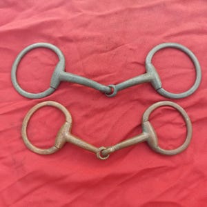 May include: Two horse bits with circular rings and a central jointed mouthpiece. The top bit is a dark grey metal, while the bottom bit is a copper-toned metal. Both pieces feature a classic snaffle design with smooth, rounded metal surfaces.