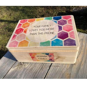 May include: A wooden box with a colorful honeycomb pattern on the lid. The lid has the text "Your family loves you more than this phone". The box has a label on the front that says "Molnár Family".