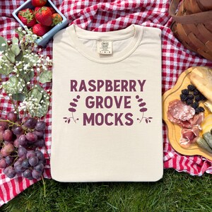 Pu&ograve; includere: Una maglietta beige con la scritta "Raspberry Grove Mocks" stampata in marrone sul davanti. La maglietta &egrave; appoggiata su una tovaglia da picnic a quadri rossi e bianchi.