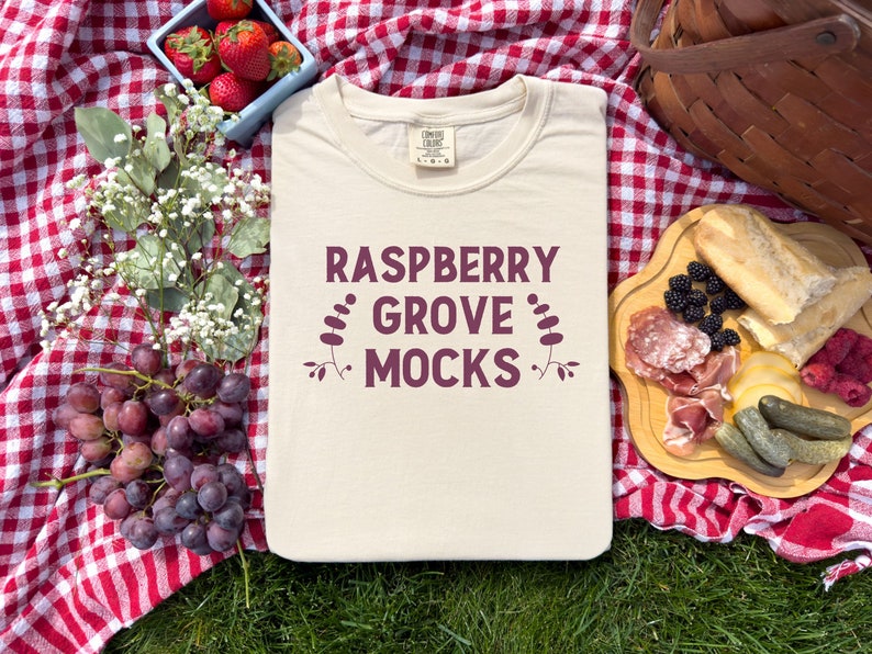 Pu&ograve; includere: Una maglietta beige con la scritta "Raspberry Grove Mocks" stampata in marrone sul davanti. La maglietta &egrave; appoggiata su una tovaglia da picnic a quadri rossi e bianchi.