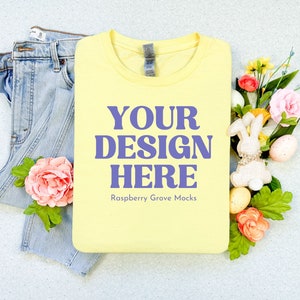 Könnte beinhalten: Ein hellgelbes T-Shirt mit dem lila Text "YOUR DESIGN HERE", neben einer hellblauen Jeans und floralen Dekorationen. Das T-Shirt ist ein Mock-up für Designzwecke. Osterei-Dekorationen, einschließlich eines Hasen, sind ebenfalls vorhanden.