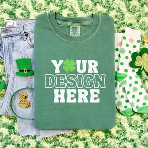 May include: A green t-shirt with the text "YOUR DESIGN HERE" in white. The t-shirt is laying on a green and white patterned surface with a pair of white socks with green shamrocks, a pair of blue jeans, and a headband with gold coins.
