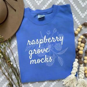 May include: A blue t-shirt with a white graphic of a raspberry bush and the text "raspberry grove mocks".