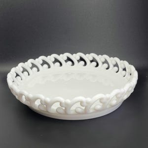 May include: A white, oval-shaped decorative bowl with a pierced, openwork rim. The bowl has a smooth, glossy surface and a slightly raised base. The rim features a repeating pattern of curved shapes.