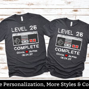 May include: Two dark gray t-shirts with a retro video game controller graphic. The text on the shirts reads "Level 26 Complete" with a heart and the date "06.04.97".
