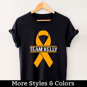 Puede incluir: Camiseta negra con una cinta naranja y el texto "TEAM KELLY" impreso en ella.
