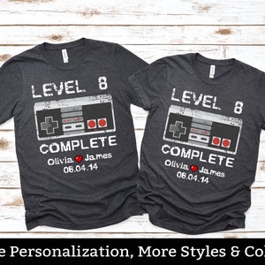 May include: Two dark gray t-shirts with the text "LEVEL 8 COMPLETE" and a retro video game controller graphic. The shirts also include the names "Olivia & James" and the date "06.04.14".