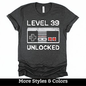 May include: A dark gray t-shirt with a white graphic of a vintage video game controller and the text "LEVEL 39 UNLOCKED".