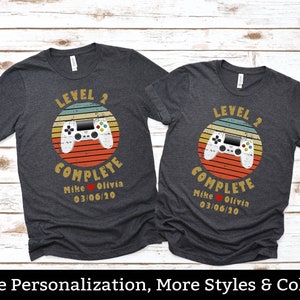 May include: Two dark grey t-shirts with a retro video game controller graphic and the text "Level 2 Complete" with a heart and names "Mike Olivia" and the date "03/06/20".