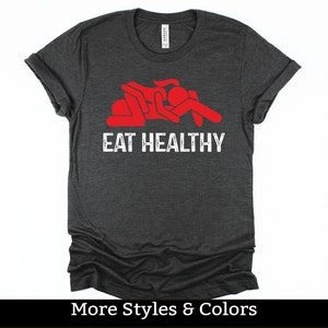 Peut inclure: Un t-shirt gris foncé avec un graphique rouge de deux personnages en bâton dans une pose suggestive et le texte "Eat Healthy".