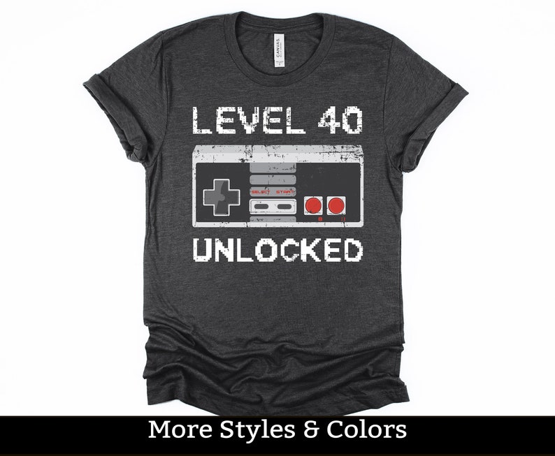 May include: Dark gray t-shirt with the text "LEVEL 40 UNLOCKED" above a pixelated video game controller graphic. The shirt also has the text "MORE STYLES & COLORS" at the bottom.