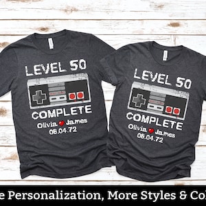 May include: Two dark grey t-shirts with a retro video game controller graphic and the text "Level 50 Complete". The shirts are personalized with the names "Olivia" and "James" and the date "06.04.72".