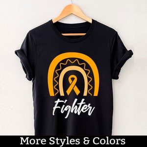 Puede incluir: Camiseta negra con una cinta naranja y un diseño de arcoíris. El texto "Fighter" está impreso en blanco debajo del diseño.