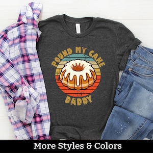 Puede incluir: Una camiseta gris oscuro con un gráfico de estilo vintage de un pastel de libra y el texto "Pound My Cake Daddy".