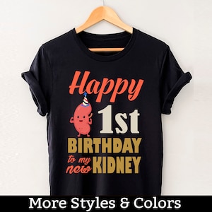 Pode incluir: Camiseta preta com um rim de desenho animado vermelho e branco usando um chapéu de festa. O texto "Happy 1st Birthday to my new Kidney" está impresso na camiseta.