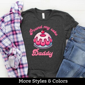 Puede incluir: Una camiseta gris oscuro con un gráfico de pastel rosa y blanco y el texto "Pound my cake Daddy".