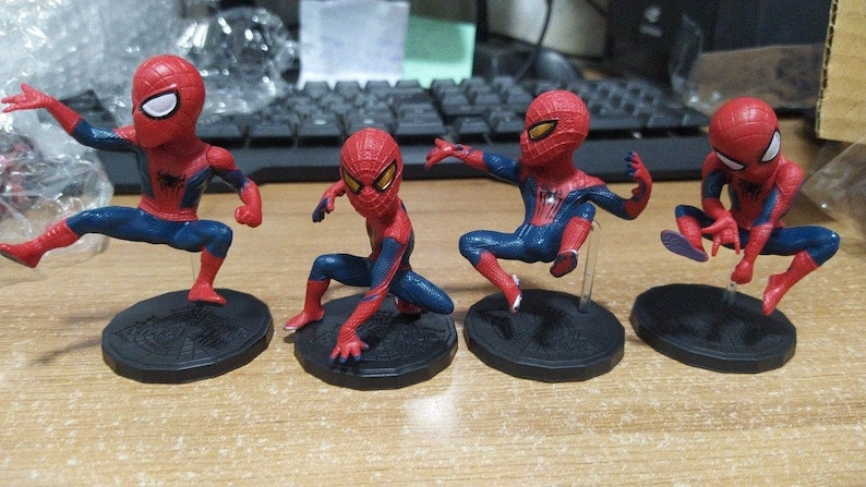 Peut inclure: Quatre figurines de Spider-Man de petite taille, rouges et bleues, avec des yeux noirs et un symbole d'araign&eacute;e blanc sur la poitrine. Chaque figurine est pos&eacute;e diff&eacute;remment et se tient sur une base noire avec un motif de toile d'araign&eacute;e.