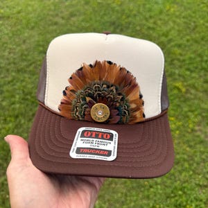 May include: A brown and cream trucker hat featuring a fan of brown, tan, and iridescent green feathers centered on the front panel. A gold-colored 12 gauge Federal shotgun shell casing is attached to the center of the feathers. An Otto brand sticker is on the brim.