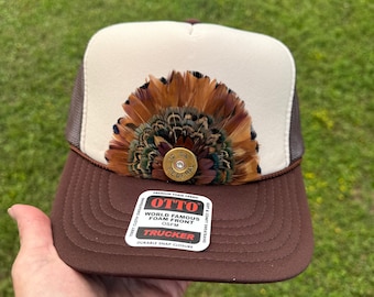 Pode incluir: Um boné trucker castanho e creme com um leque de penas castanhas, bege e verde iridescente no painel frontal. Um invólucro de cartucho de caçadeira Federal de calibre 12 dourado está fixado no centro das penas. Um autocolante Otto está na pala.