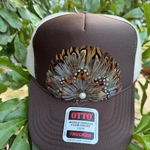 May include: A brown trucker hat with a white mesh back and a foam front panel. The front features a decorative fan-shaped arrangement of brown, tan, and grey feathers centered with a small white pearl. A sticker on the brim reads OTTO TRUCKER.