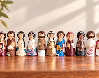 May include: A set of fourteen hand-painted wooden peg dolls depicting various religious figures. Each figure features distinct clothing, colors, and accessories, including floral patterns, robes, and symbols, arranged in a single horizontal row.