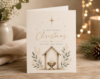 May include: A cream-colored greeting card featuring a gold foil star at the top and the text A VERY MERRY Christmas TO YOU AND YOURS. Below the text is a watercolor illustration of a manger scene inside a wooden stable with green foliage.