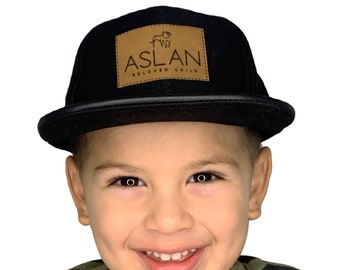 May include: A young boy wearing a black and brown baseball cap with a leather patch that says "ASLAN BELOVED CHILD".