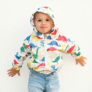May include: A white hooded jacket with a colorful dinosaur print. The jacket has a zipper closure and is made of a lightweight fabric.